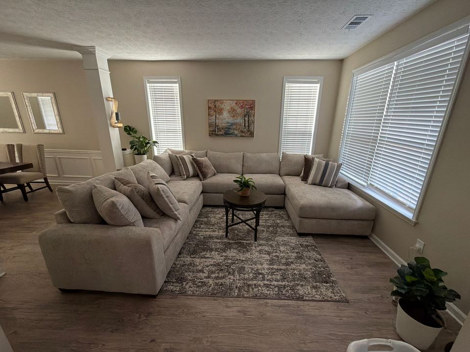 Living room with a large beige sectional sofa, a patterned area rug and white window blinds